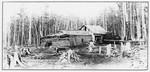 Black-and-white historical photograph of a small log cabin situated beside a narrow stream or millrace in a forested clearing. The cabin has a low-pitched roof and a stone chimney, with a simple wooden structure extending from one side. A rough dirt path and wooden footbridge cross the water in the foreground. Tall leafless trees surround the site, creating a secluded woodland setting. The image has a vintage appearance and is framed by a decorative border.